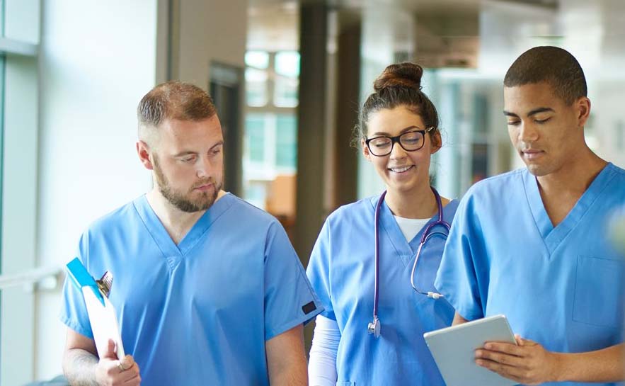 Three healthcare professionals in blue scrubs walking through a medical facility, discussing information on a digital tablet.