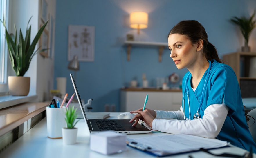 female healthcare worker in blue scrubs sitting at her laptop