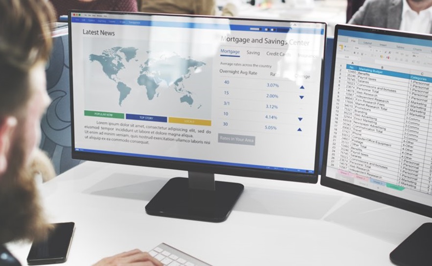 Person working at a desk with two computer monitors displaying a world map and latest news headlines, and a spreadsheet with mortgage and savings data.
