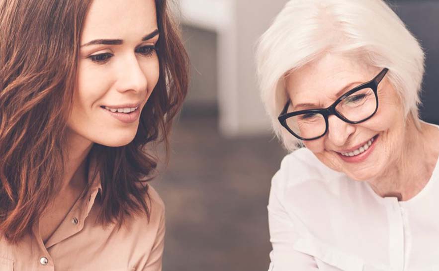Younger woman with brown hair in a beige shirt and older woman with white hair, glasses, and a white shirt, sitting closely together.