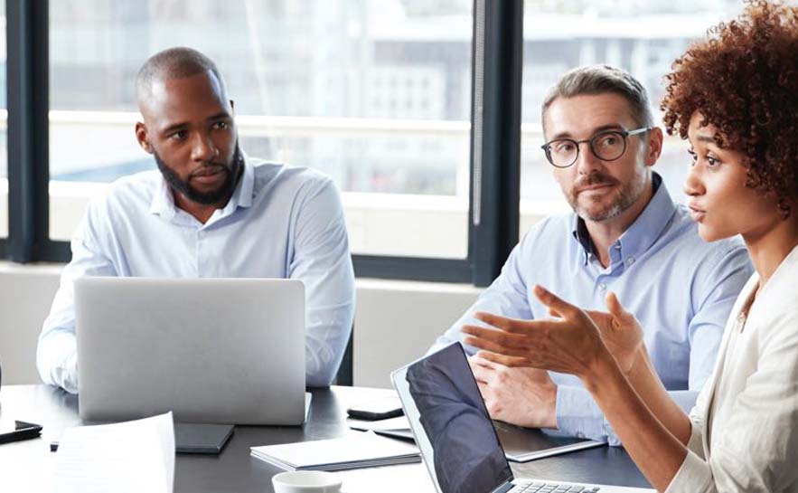 Three people sitting at a table in an office setting, engaged in a discussion with laptops and documents on the table.