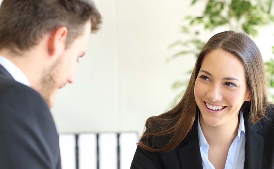 Two people in a professional setting having a conversation. One is a woman wearing a blazer over a light blue shirt, while the other person is a man and is also dressed in business attire.