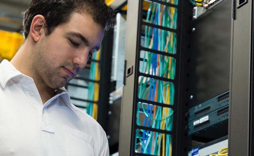 Man wearing a white shirt standing next to a computer server with various colored cables and network equipment.
