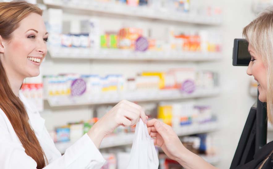Two women in a pharmacy exchanging a bag, with shelves of various medications and products in the background.
