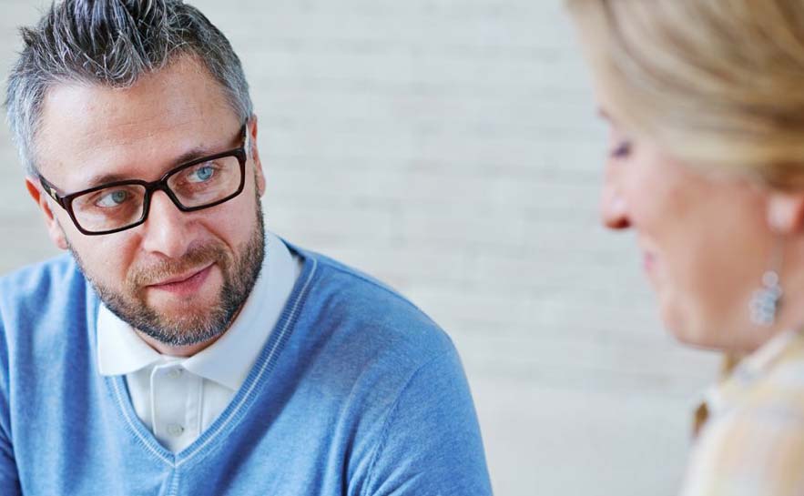 Two people sitting and talking. The man has grey hair, glasses and a sweater.