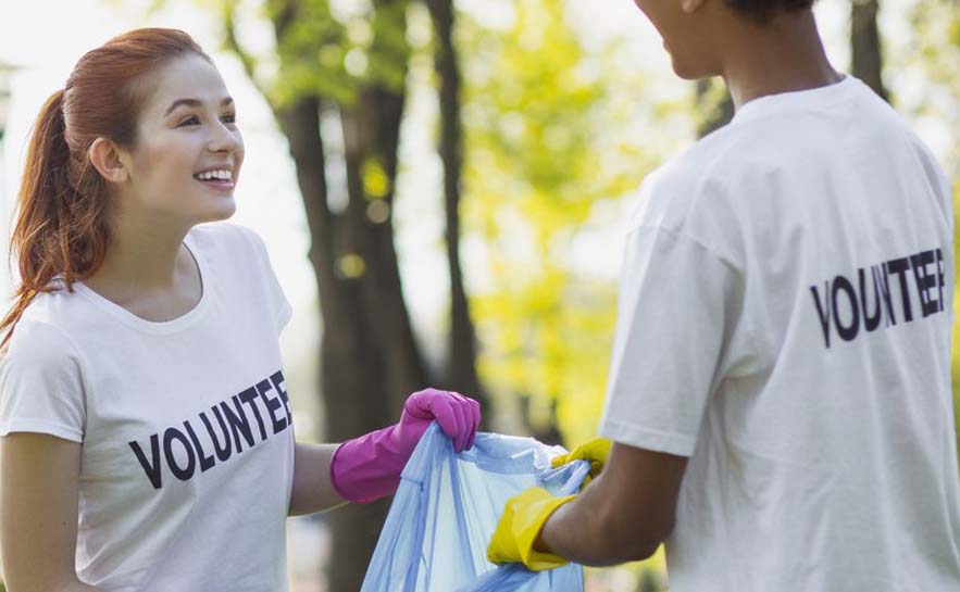 Two people wearing white t-shirts with the word 'VOLUNTEER' printed on them, holding a blue trash bag and wearing rubber gloves while cleaning up outdoors.