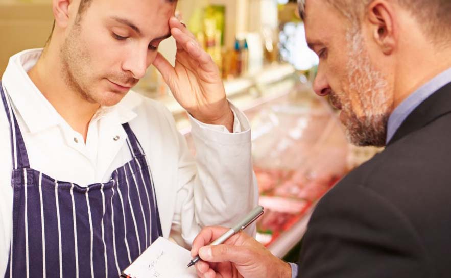 A person in an apron talking to another person in a suit who is holding a pen and notepad. They appear to be in store or market.