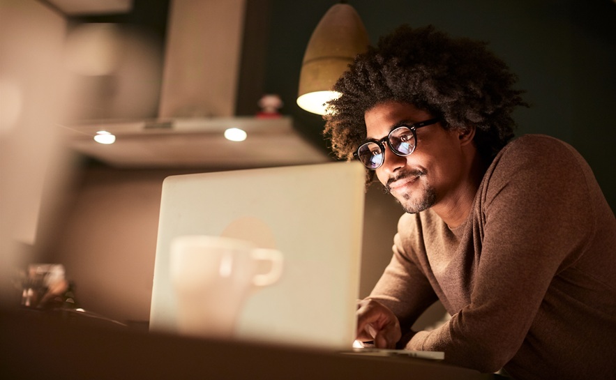 african american male wearing a long sleeve shirt and glasses sitting at a desk looking at his computer