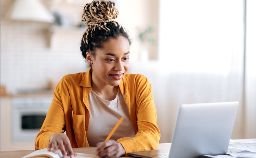 african american woman looking at her laptop writing notes in a notebook wearing an orange long sleeve shirt and white tshirt