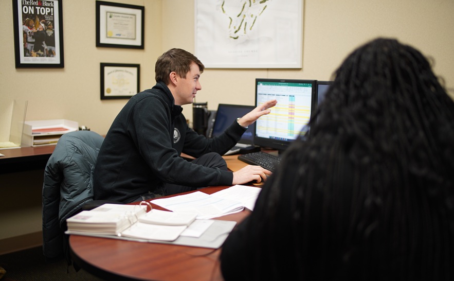 south university financial planner in a dark shirt is pointing at his computer monitor showing something to another person sitting at the desk