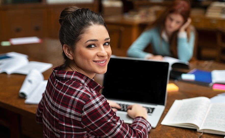 young lady wearing a plaid shirt typing on a laptop in a library with several books surrounding her